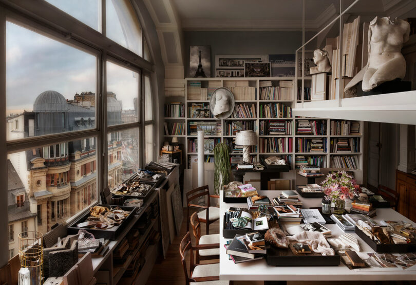 Art studio with large window overlooking cityscape. Shelves filled with books and sculptures, tables covered with art materials, and a vase of flowers on a central table.