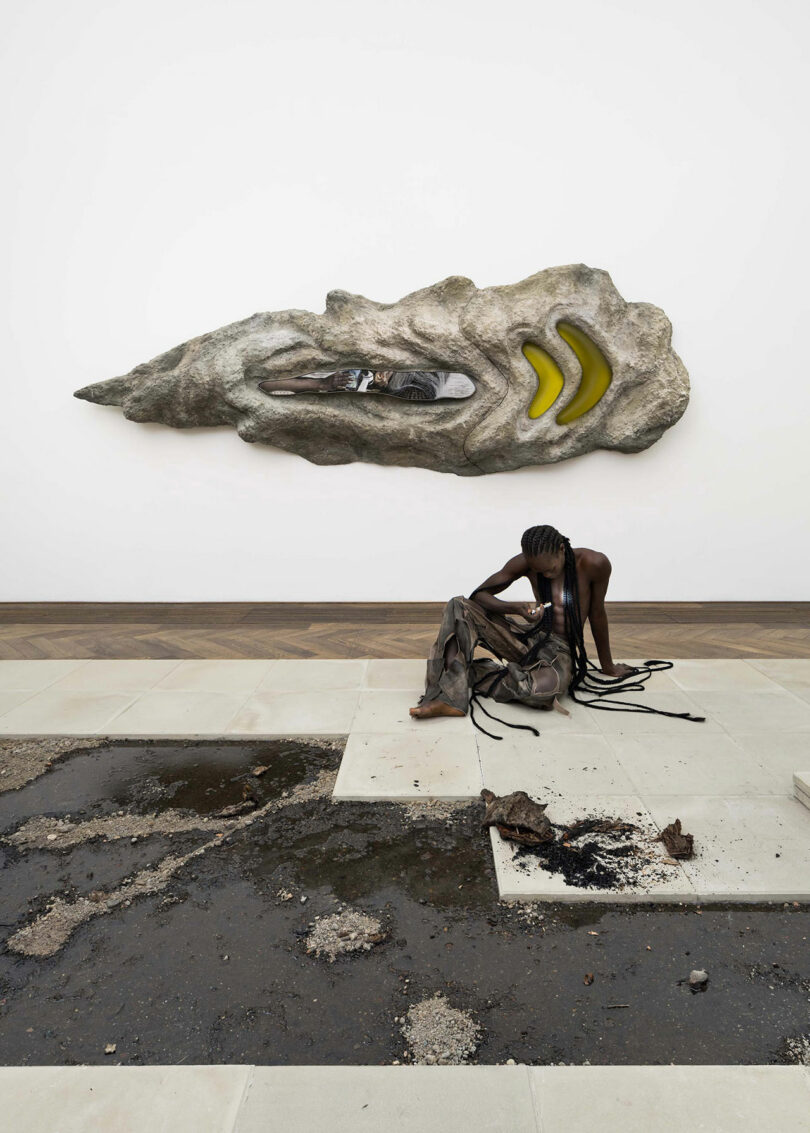 A person with long braids sits on the floor surrounded by ropes, with a large abstract sculpture mounted on the wall behind them. Some debris and water are on the floor.