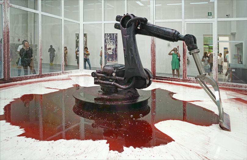 A mechanical arm continuously scrapes a pool of dark red liquid in a glass-walled room while onlookers observe from outside.