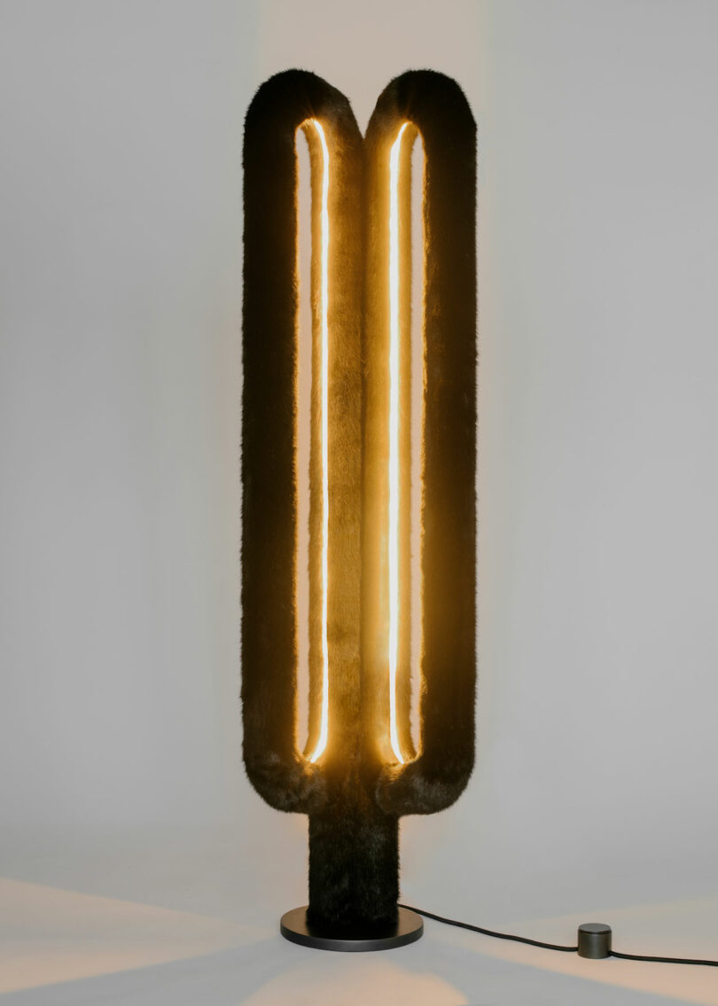 Tall, fur-covered floor lamp with two vertical glowing light tubes, standing on a round base, plugged into a wall.