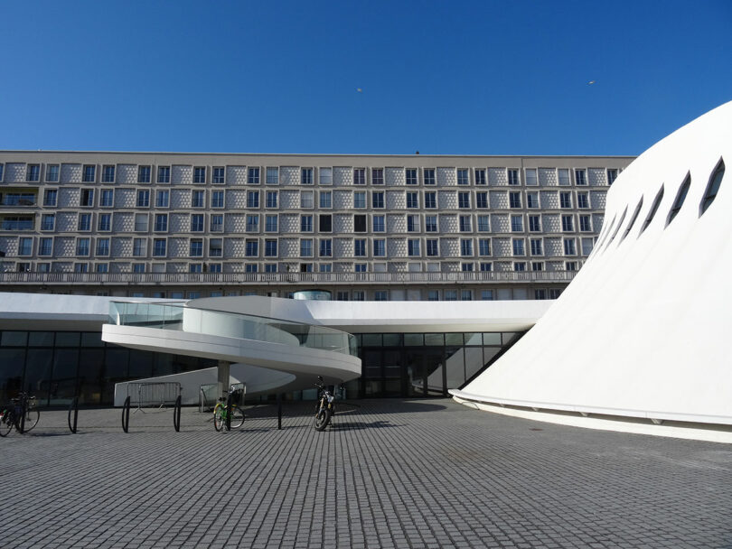 Modern architectural building with a spiral walkway, bicycles parked nearby, and a tall structure in the background, set against a clear blue sky.