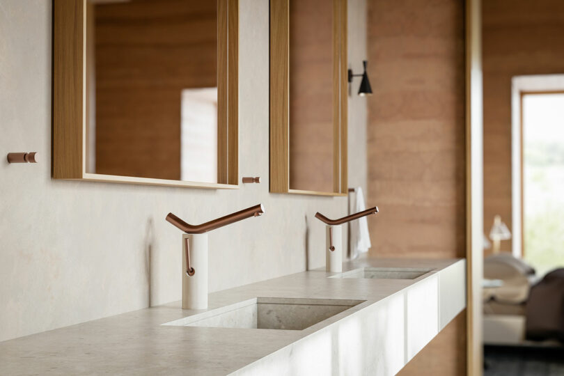 Modern bathroom with two sinks and sleek, angled faucets on a light-colored countertop. Wooden frames surround the mirrors, and the walls have a warm, textured finish. Natural light filters in.