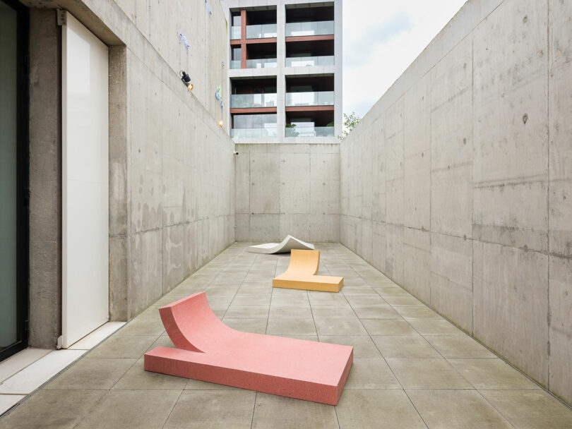 Minimalist concrete courtyard with three colorful, abstract benches in red, yellow, and white. A modern building is visible in the background