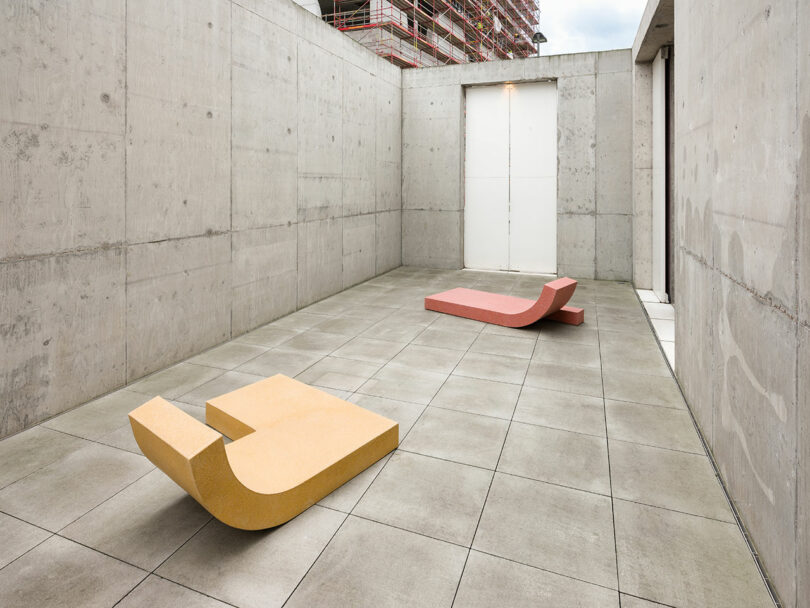 Concrete courtyard with two minimalist, curved benches in yellow and red, set against a modern, industrial backdrop