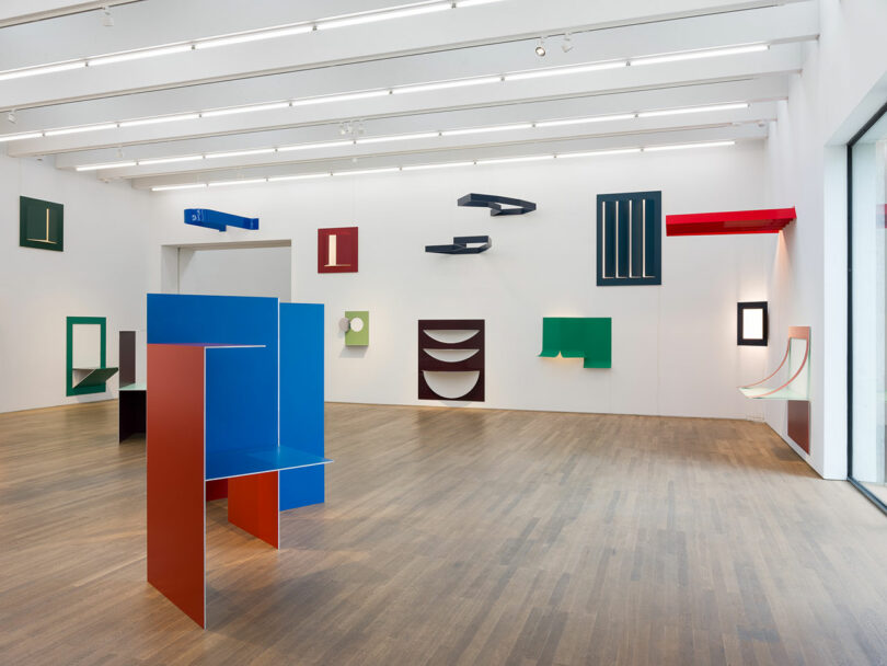 Art gallery with abstract geometric sculptures and wall-mounted pieces in various colors, displayed in a spacious room with wooden floors and white walls