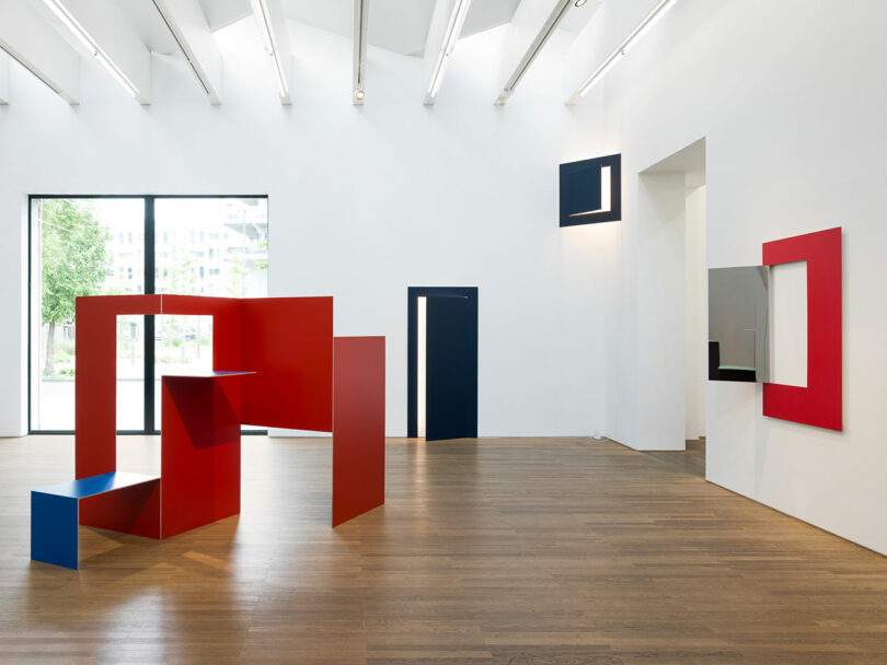 Modern art gallery with geometric sculptures and installations in red, blue, and black on wood flooring and white walls