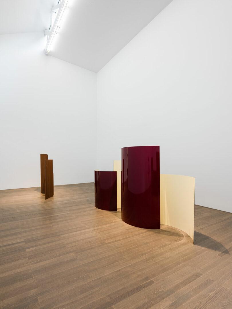 An art gallery room with modern sculptures featuring curved panels in burgundy and beige, placed on a wooden floor