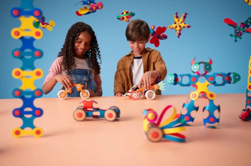 Two children play with colorful toy constructions on a table, with more toys floating in the background against a blue backdrop