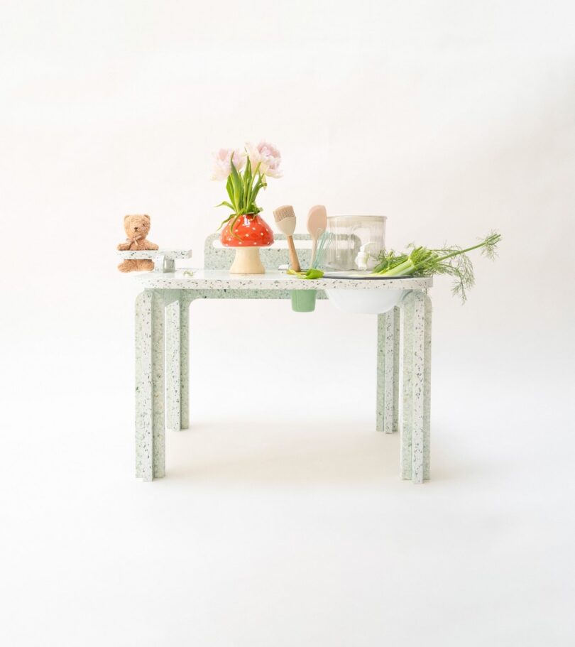 A minimalist table with terrazzo design holds a potted plant, a ceramic mushroom-shaped item, wooden utensils, herbs, and a jar, set against a plain background