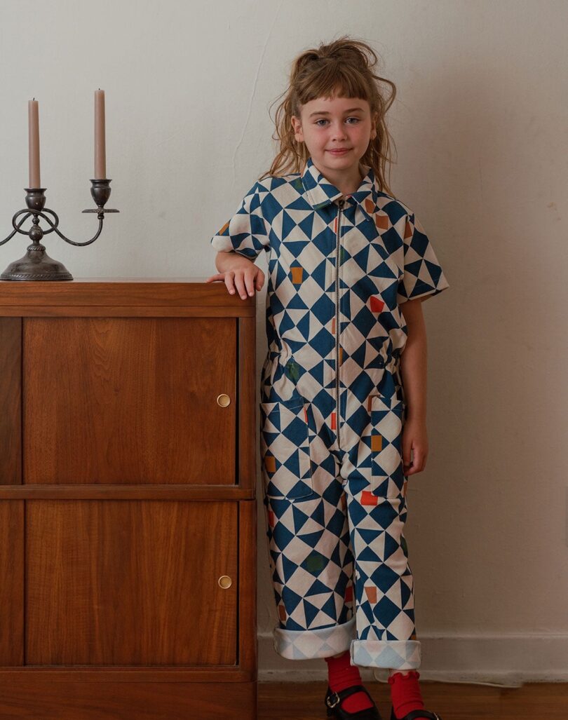 Child in a geometric-patterned jumpsuit stands beside a wooden cabinet with unlit candles against a plain wall