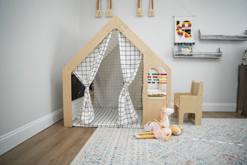 Children's playroom with a house-shaped tent, small wooden chair, abacus, and plush toys on the floor. Colorful wall decorations hang nearby