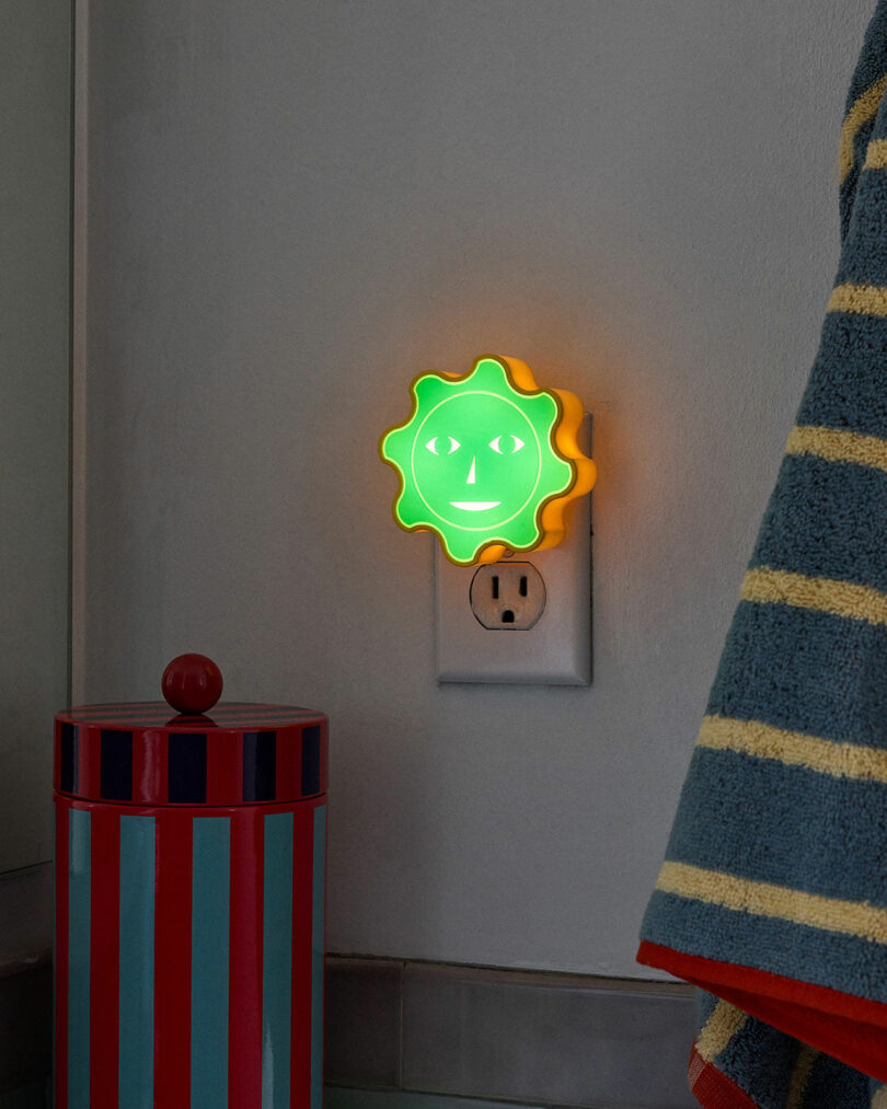 Modern kids gifts include this vibrant sun-shaped nightlight with a cheerful face, snugly plugged into a wall socket beside a striped container.