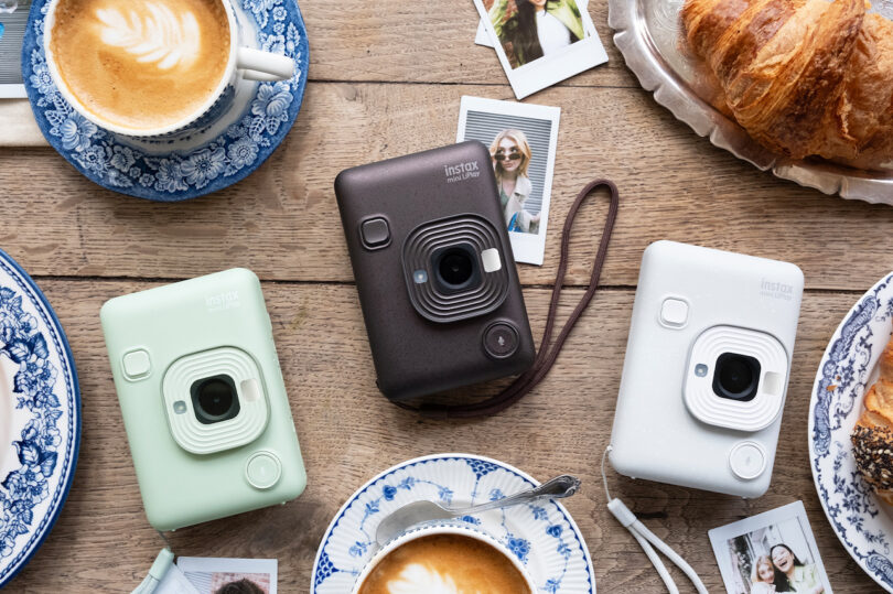 3 instax Mini LiPlays in Matcha Green, Deep Bronze, and Misty White on a wood table surrounded by printed photos, coffees with latte art, and croissants