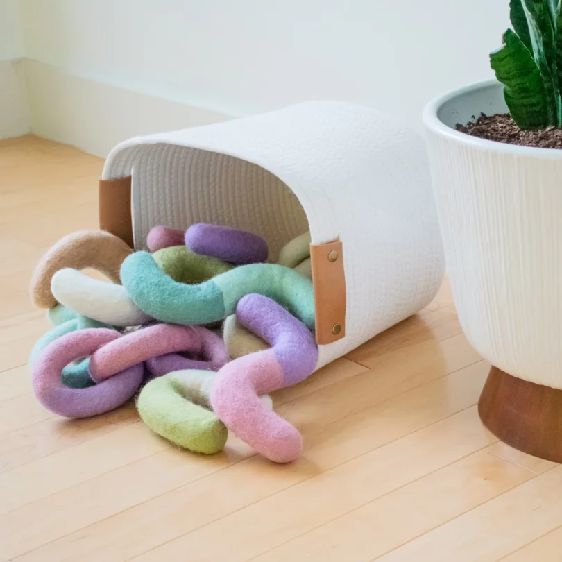 Various felt toys for dogs outside of a tipped-over white basket with light brown leather handles