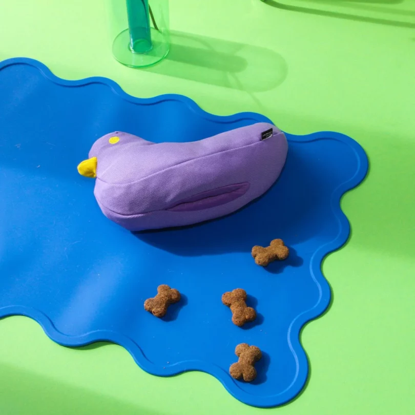 A purple bird dog toy tipped over exposing a slit on the bottom meant for treats, with four treats next to it; the toy is on a blue mat, which is on a lime green floor