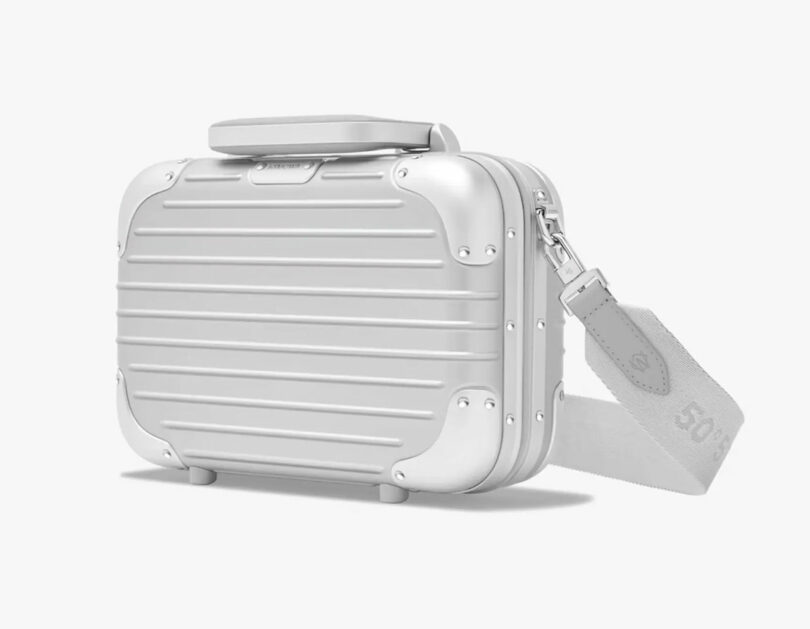 A small silver hard-shell suitcase with a handle on top and a detachable shoulder strap, angled on a white background.