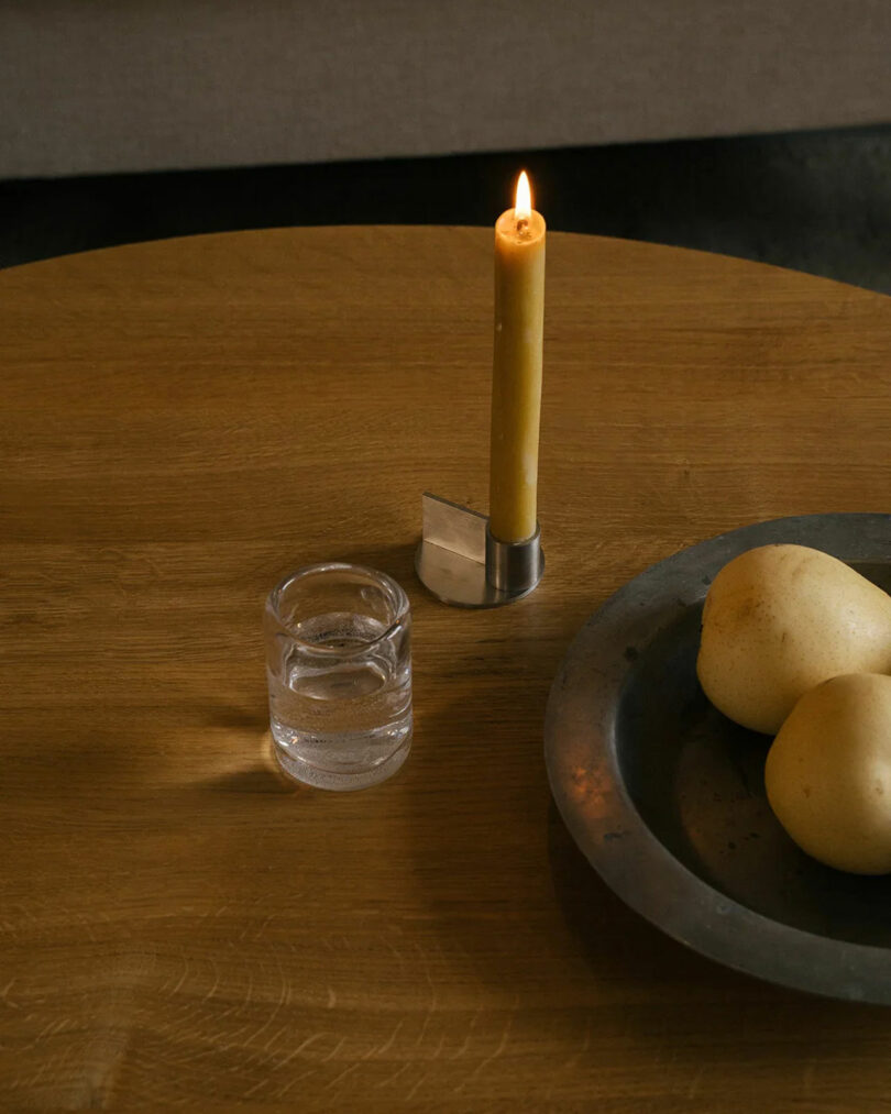 A lit candle in a holder, a glass of water, and a metal dish with two pears on a wooden table.