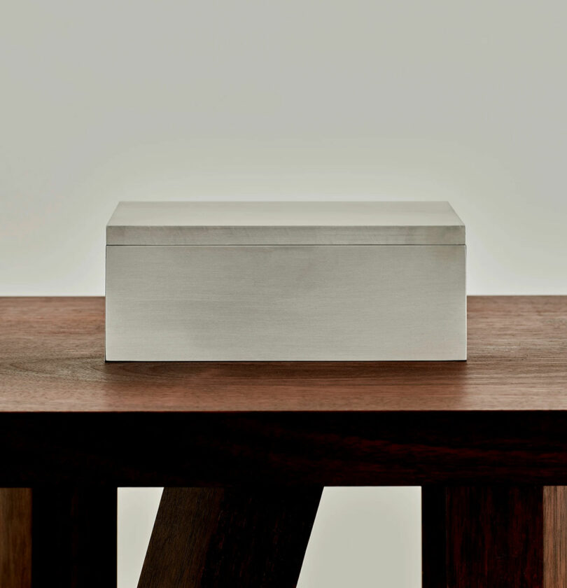 A closed, rectangular silver box sits on a wooden surface against a neutral background.