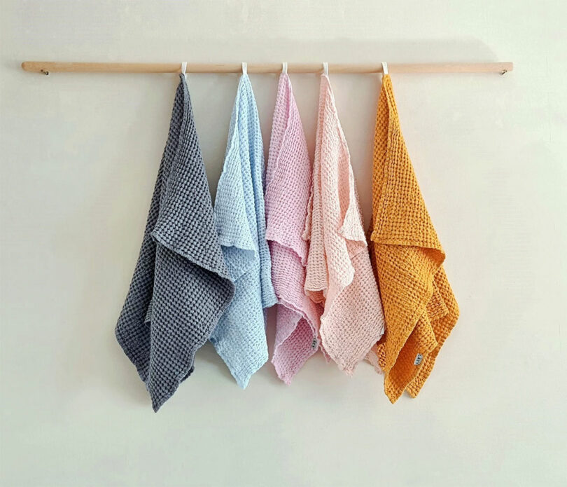 Five waffle-textured towels in gray, light blue, pink, peach, and mustard hang on a wooden rod against a white wall.