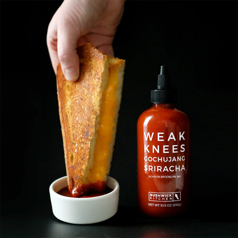 Hand dipping a grilled cheese sandwich into a sauce, next to a bottle of "Weak Knees Gochujang Sriracha" sauce from Bushwick Kitchen.