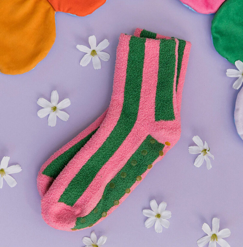 Pink and green striped socks with non-slip grips are placed on a purple surface surrounded by small white flowers.