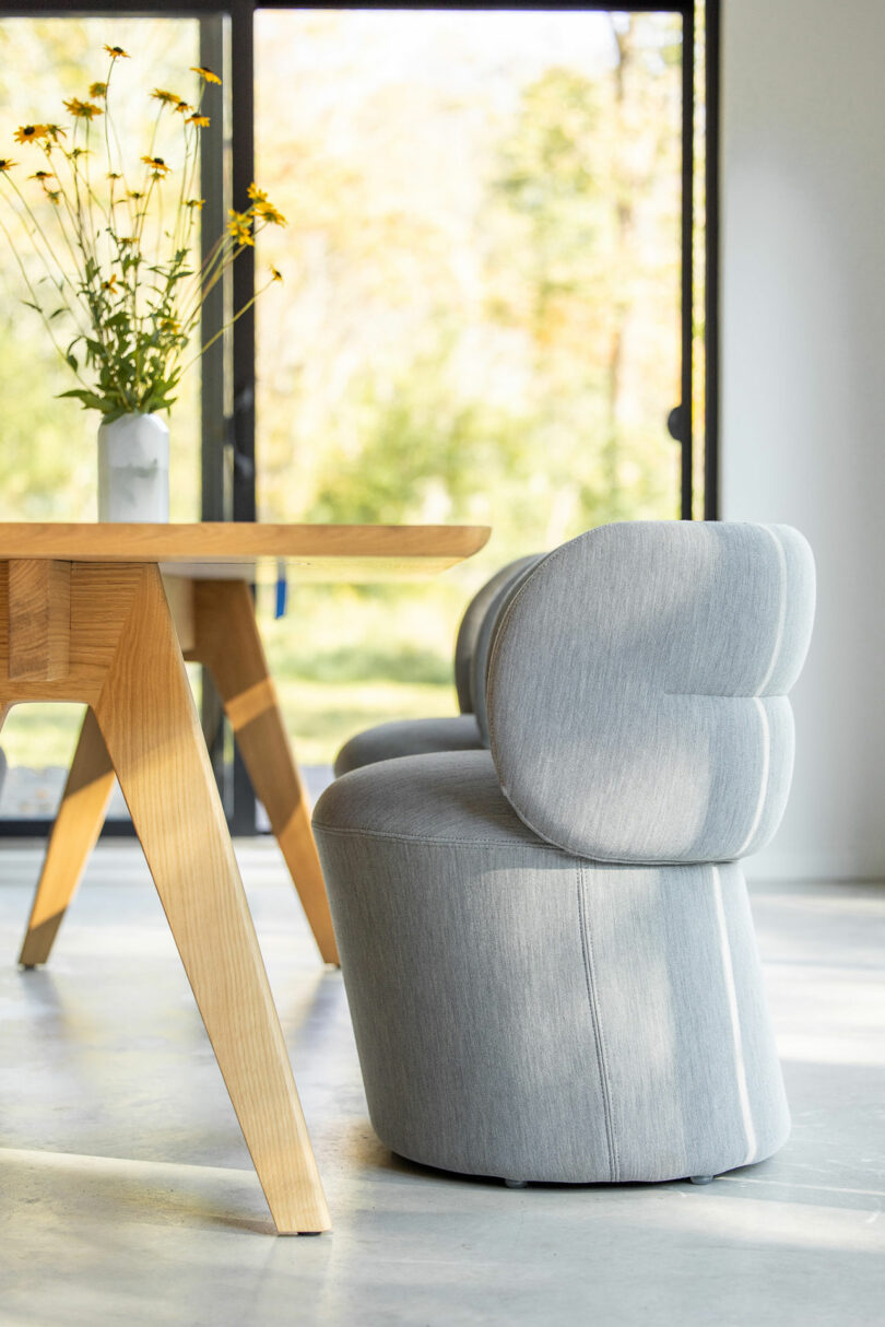 A gray upholstered chair next to a wooden table with a vase of yellow flowers, set against a backdrop of large windows and greenery outside.