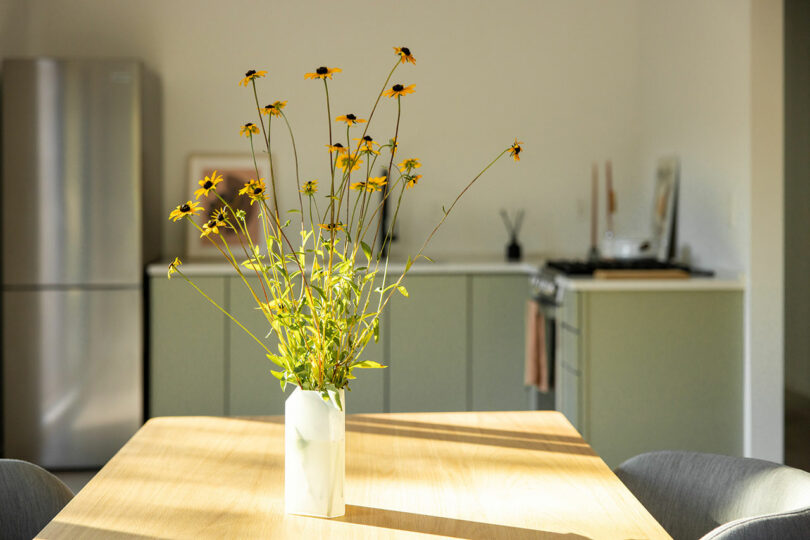 A vase with yellow flowers is centered on a light wooden dining table. In the background, there is a modern kitchen with a refrigerator and muted green cabinets.