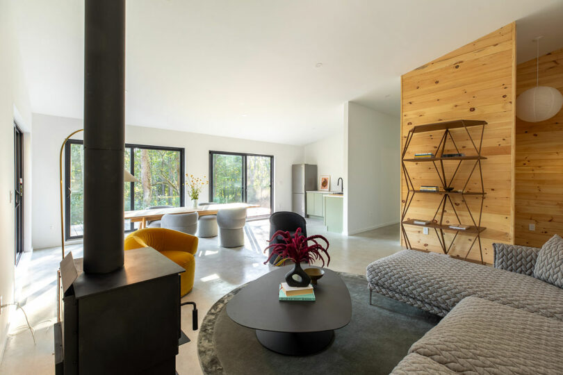 Modern living room with a wood stove, gray sectional sofa, round coffee table, yellow chair, and a wooden open-shelf bookcase. Large windows overlook a wooded area.