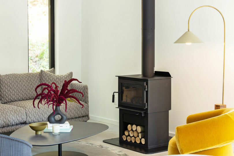 A modern living room with a wood-burning stove, a gray sofa, a yellow chair, a round table with a plant, and a standing lamp. Logs are stored under the stove.