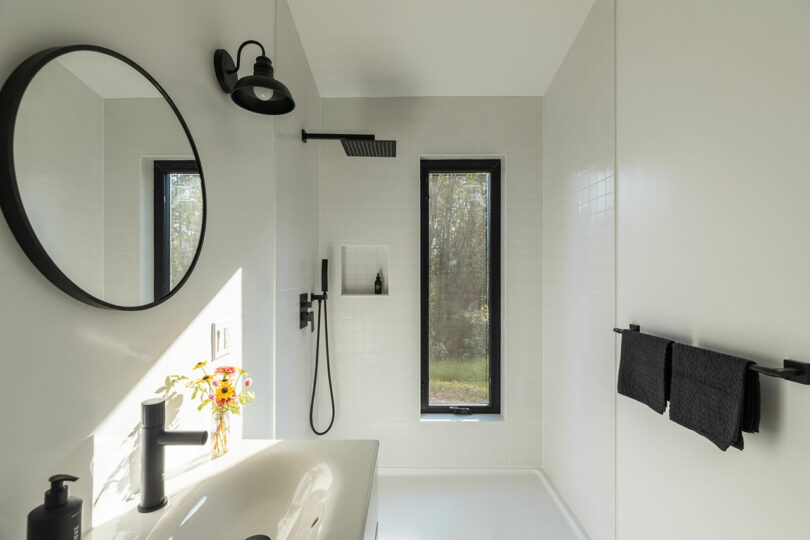 Minimalist bathroom with a white sink, black fixtures, round mirror, and a window. Sunlight illuminates a small flower vase on the counter.