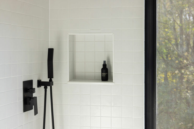 White tiled shower with a black handheld showerhead and a niche holding a single black bottle.