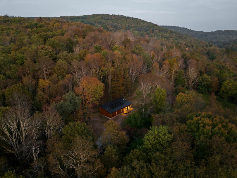 A cabin with lights on is nestled in a densely forested area with rolling hills in the background.