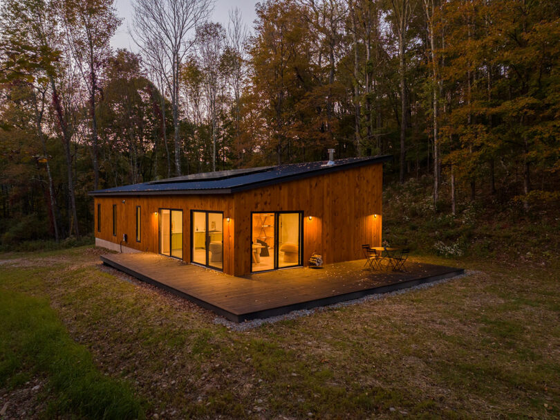 A modern wooden cabin with large windows and a wrap-around deck is surrounded by a forest. Outdoor lights illuminate the scene during twilight.