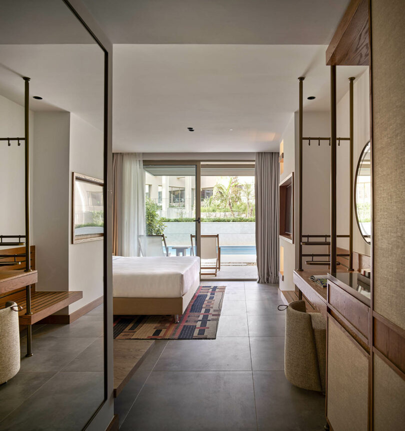 Hotel room with a bed, desk, and large mirror. Glass doors open to a poolside view with patio furniture visible outside. Neutral color palette and modern decor.