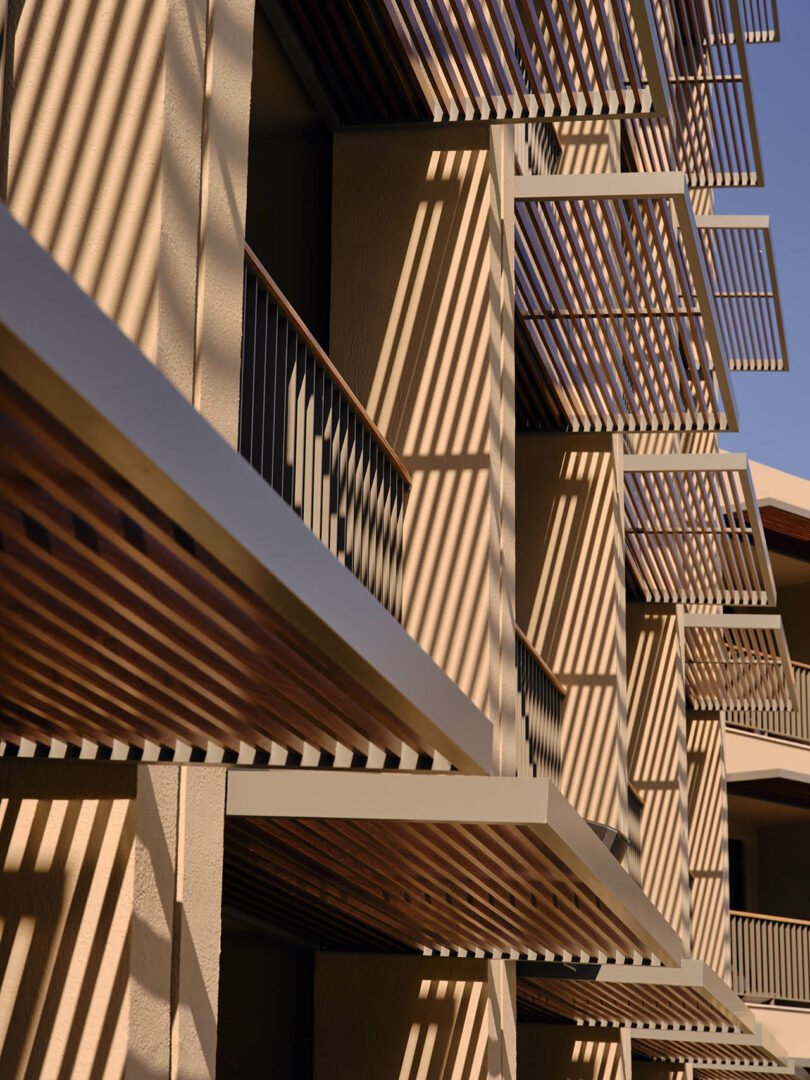 Facade of a modern building with horizontal and vertical slatted patterns creating angular shadows on beige walls, captured under direct sunlight.