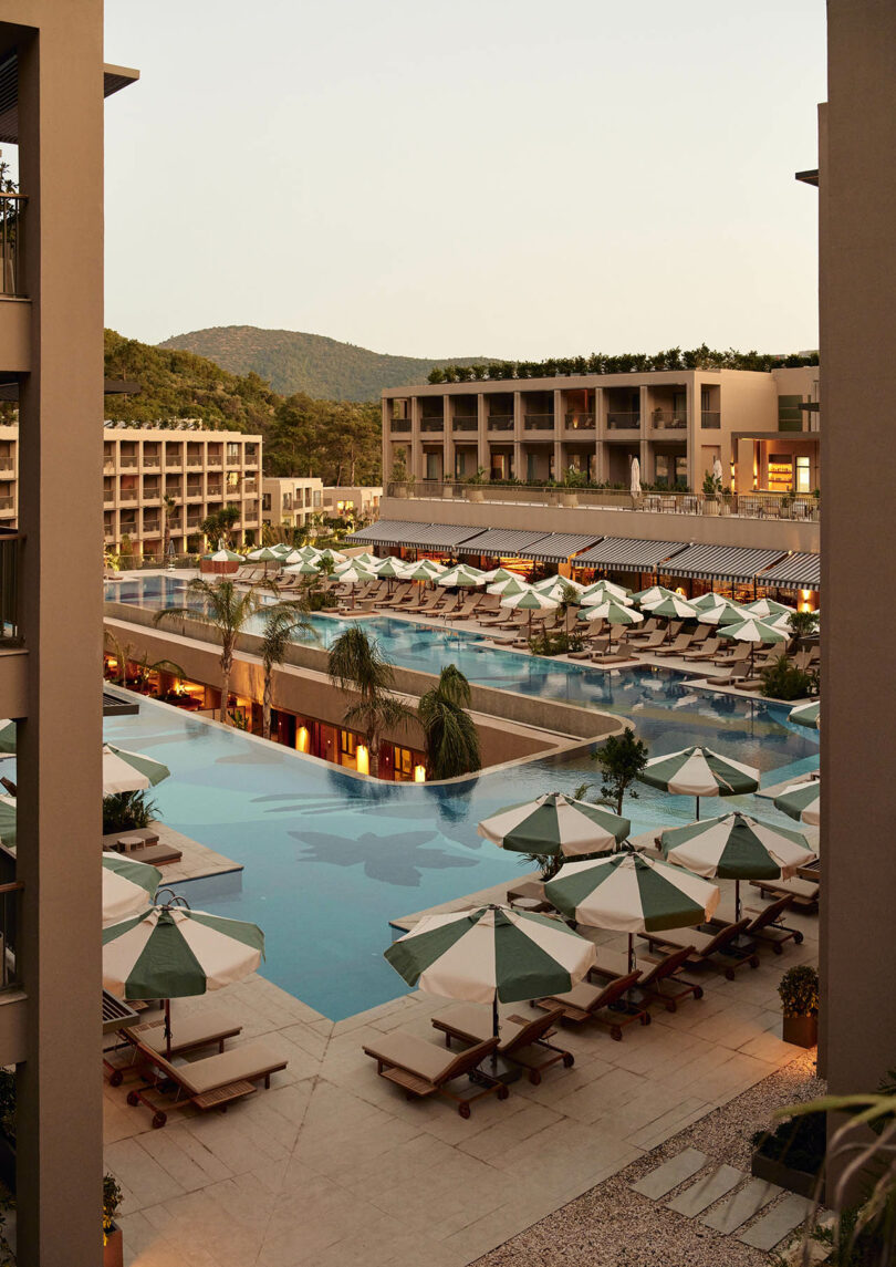 Luxury resort with multiple pools, lined with loungers and umbrellas. Surrounding modern buildings and hills are visible in the background.