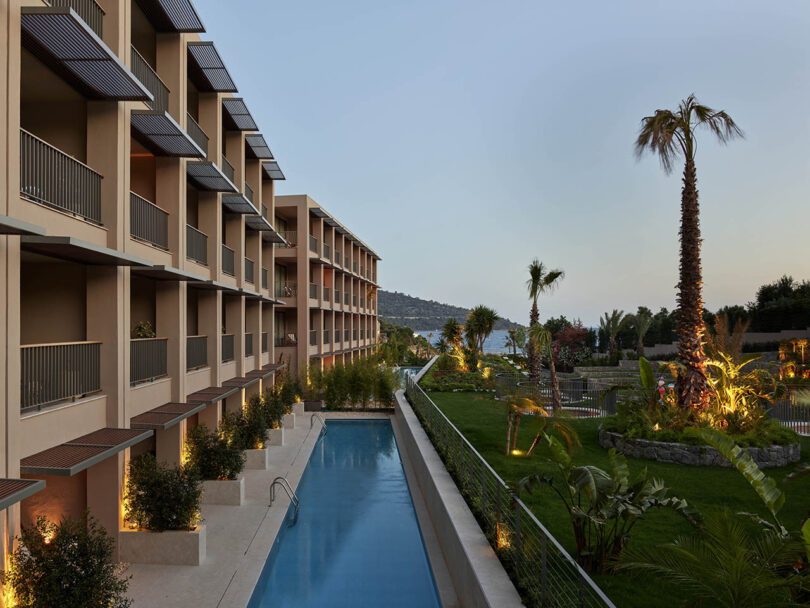 A modern hotel exterior with multiple balconies overlooks a narrow pool beside a landscaped garden with palm trees. The scene is set during dusk.