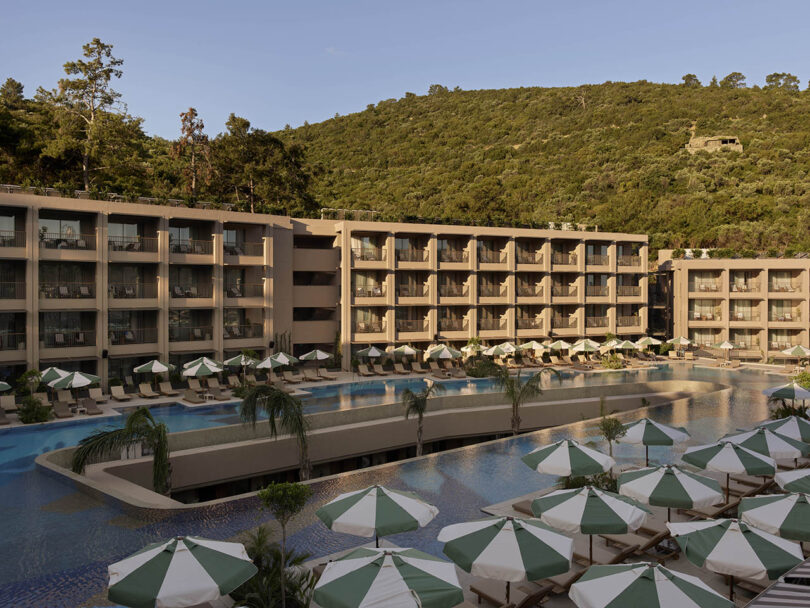 A large resort with multiple floors overlooks a pool area with green and white umbrellas. Lush green hills are visible in the background.