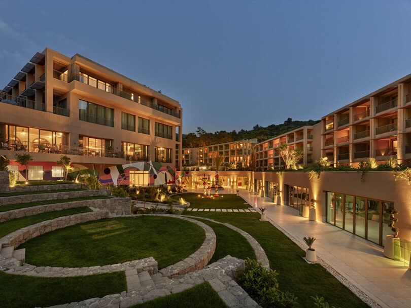 Modern hotel with illuminated windows and a courtyard featuring tiered gardens and pathways at dusk.