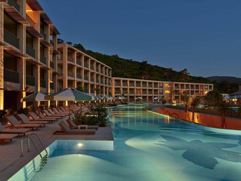 Luxury resort with modern architecture, featuring a large pool and poolside loungers, set against a backdrop of trees at dusk.