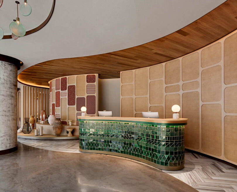 A modern hotel lobby with a curved green-tiled reception desk, patterned wall panels, and decorative vases.