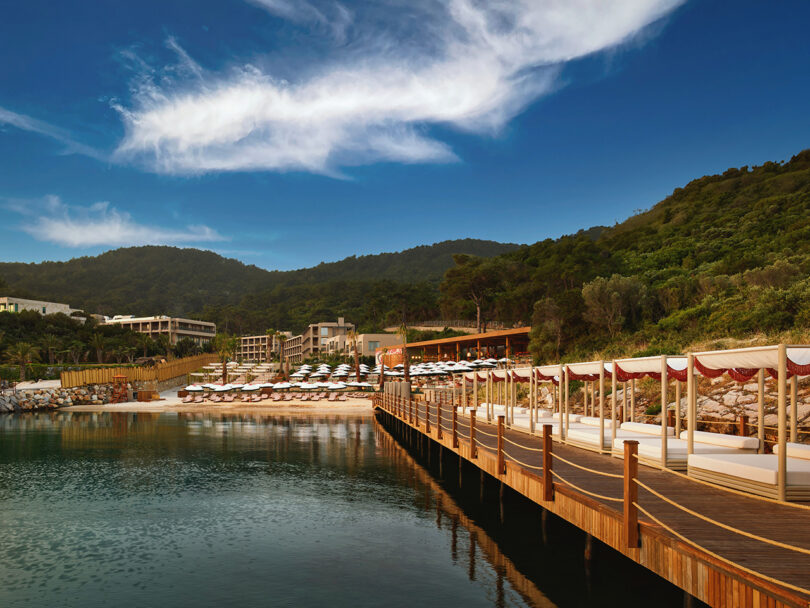 Wooden pier leading to a beach with sun loungers and cabanas, surrounded by green hills and under a blue sky with wispy clouds.