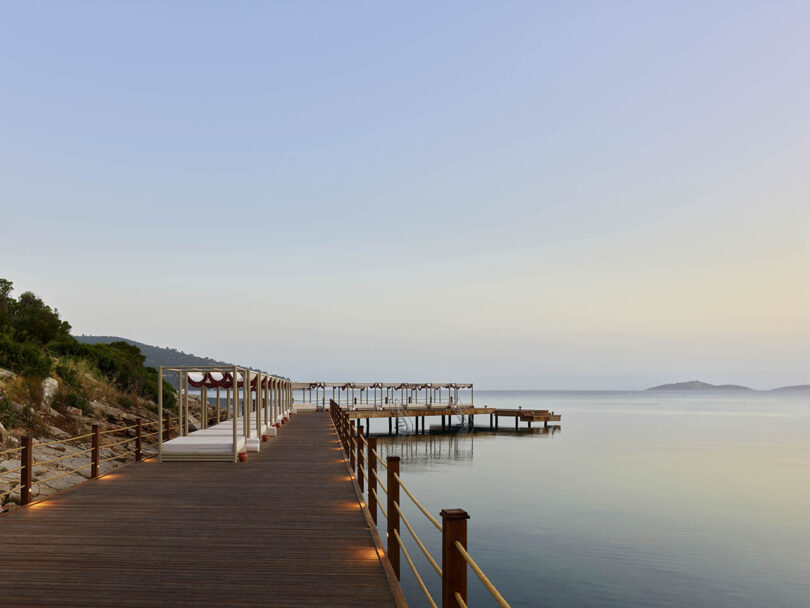 A wooden boardwalk extends over calm water with distant hills under a clear sky. A series of canopied areas are situated along the boardwalk.