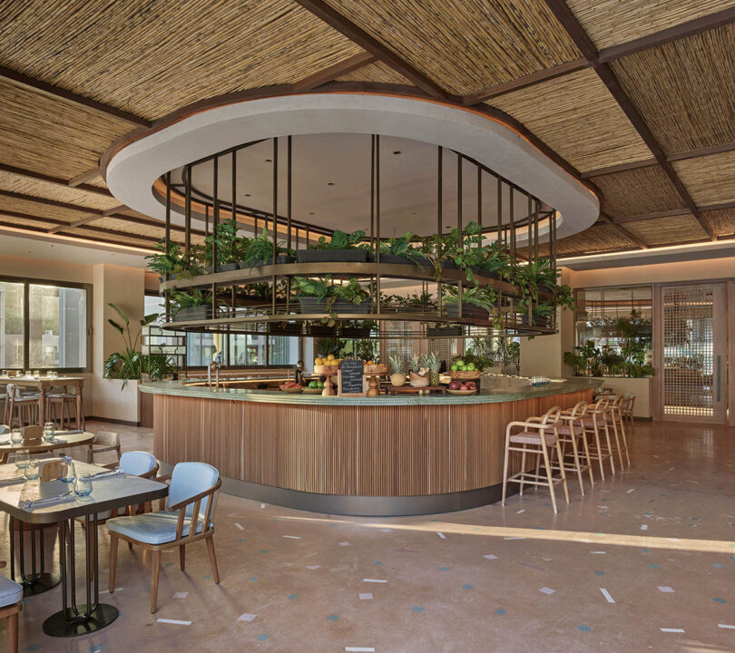 Modern restaurant interior with a central circular counter surrounded by stools. Ceiling features wood textures, and plants adorn the space. Tables and chairs are neatly arranged on a terrazzo floor.