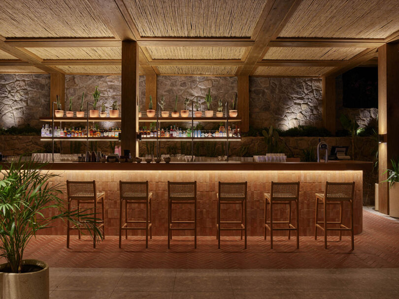 A warmly lit bar with six wicker chairs, a stone wall backdrop, and shelves stocked with various bottles and potted plants. The ceiling features wooden beams and straw accents.