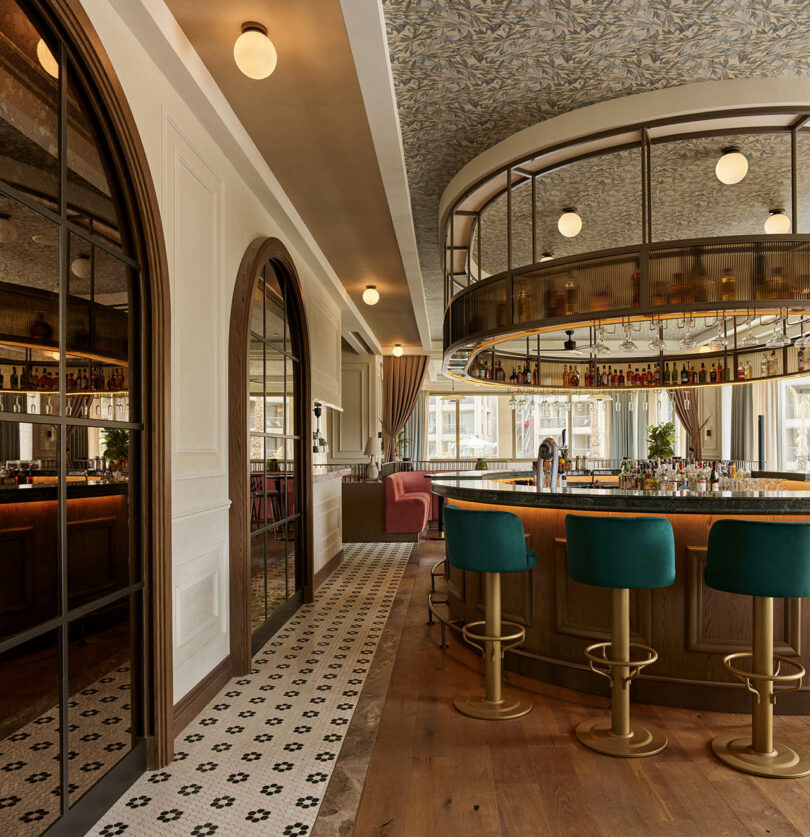 Elegant bar interior with green stools, circular counter, and curved ceiling. Patterned floor tiles and arched wooden doors enhance the stylish ambiance.