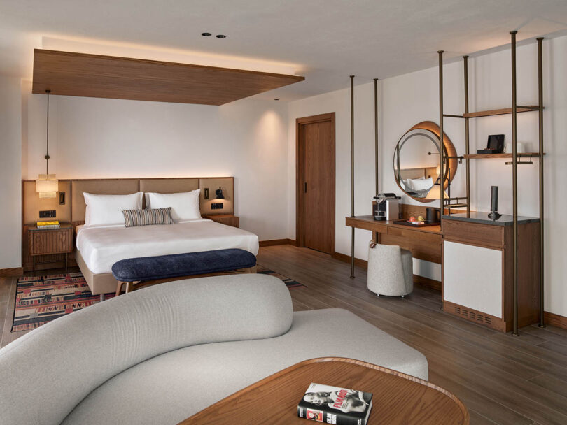 Modern hotel room with a large bed, circular mirror, desk, and seating area. Decor includes wood accents and soft lighting.