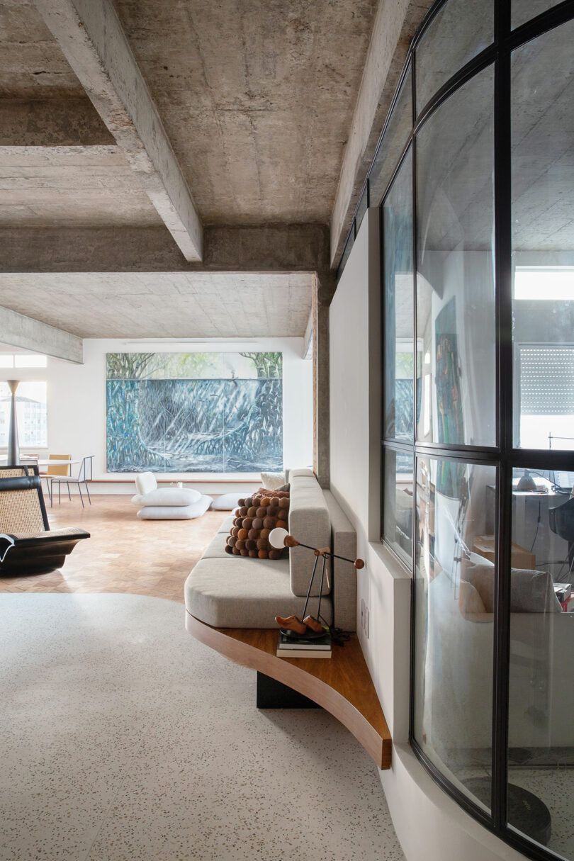 Modern living room with minimalist decor, featuring a curved sofa with round cushions, concrete ceiling, floor-to-ceiling windows, and abstract wall art in the background.