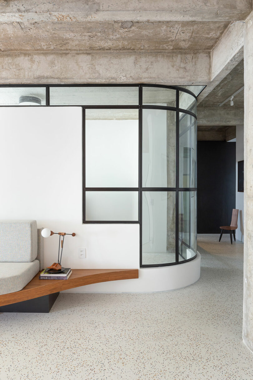 Modern interior with curved glass wall, gray and white color scheme, concrete ceiling, and a wooden bench with a small table lamp.