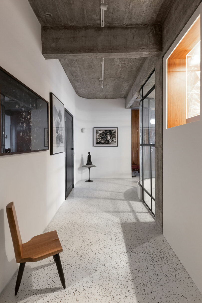 Modern hallway with concrete ceiling, white walls, and pebble-textured floor. Features a wooden chair, framed art, a small side table with a decorative bell, and a glass partition.